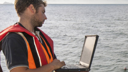 Man aan de waterkant met een computer en een oranje veiligheidsvest